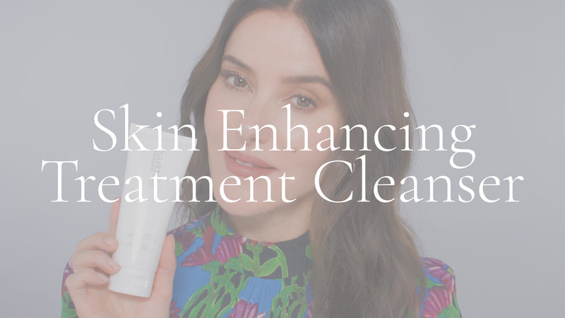 Poster image with white overlay for a video. Lisa Eldridge is in front of a grey background, holding a tube of Skin Enhancing Treatment Cleaner. On top of the image is the text, “Skin Enhancing Treatment Cleanser”.