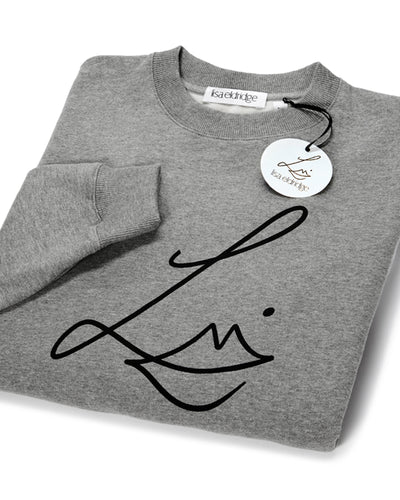 The Lisa Eldridge Studio Sweatshirt in grey folded up. The crewneck and the cuffs have a fine rib. The Lisa Eldridge logo is large on the front. There is a label that says “lisa eldridge’ sewn on the inside. Attached to the size label is a circular card tag on a black ribbon with the Lisa Eldridge logo in gold.