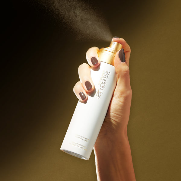 Hand pressing the spray nozzle of Seamless Skin and Makeup Enhancing Mist against a dark background