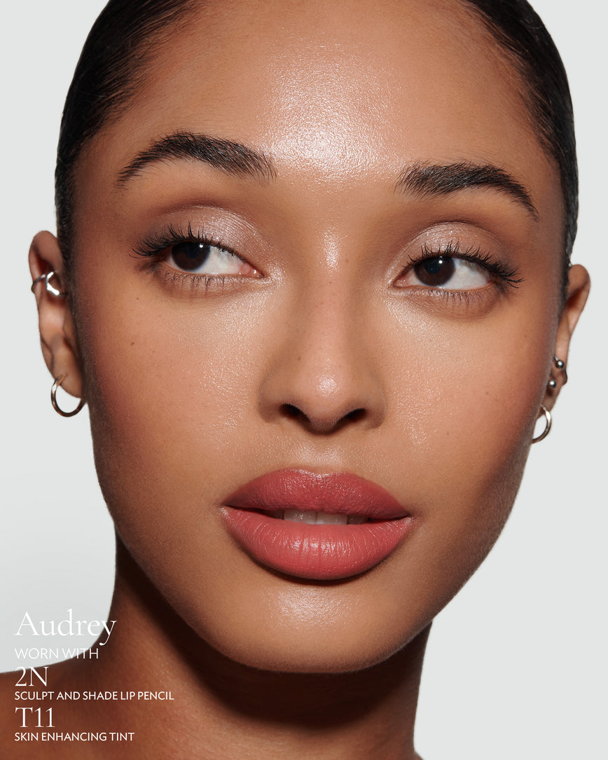 Close-up of a woman with a grey background, focusing on the lip colour - salmon pink Audrey, with notes on the 2N Sculpt and Shade Pencil and T11 Skin Enhancing Tint
