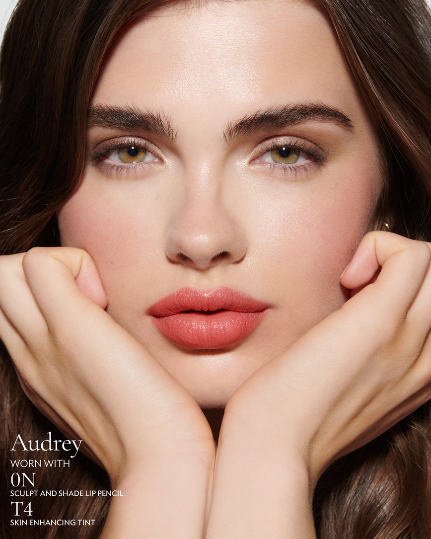 primary_meta_image Close-up of a woman with a grey background, focusing on the lip colour - salmon pink Audrey, with notes on the 0N Sculpt and Shade Pencil and T4 Skin Enhancing Tint