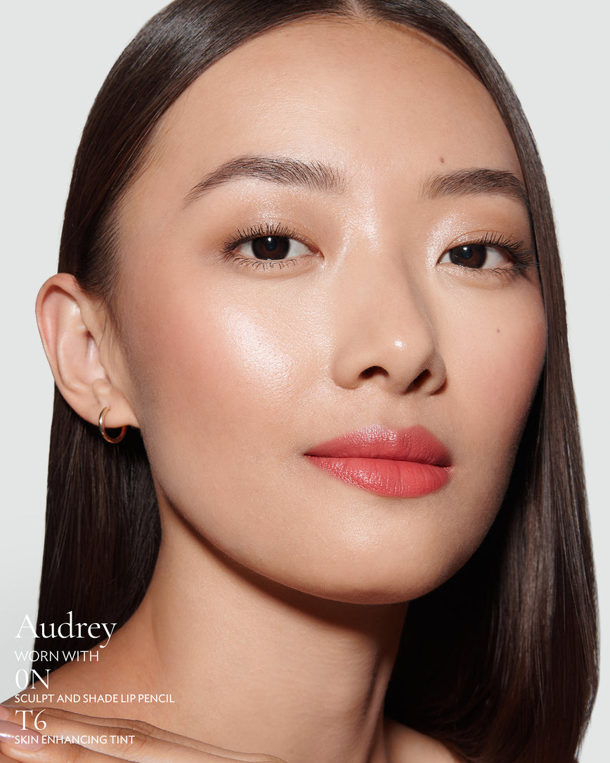 Close-up of a woman with a grey background, focusing on the lip colour - salmon pink Audrey, with notes on the 0N Sculpt and Shade Pencil and T6 Skin Enhancing Tint