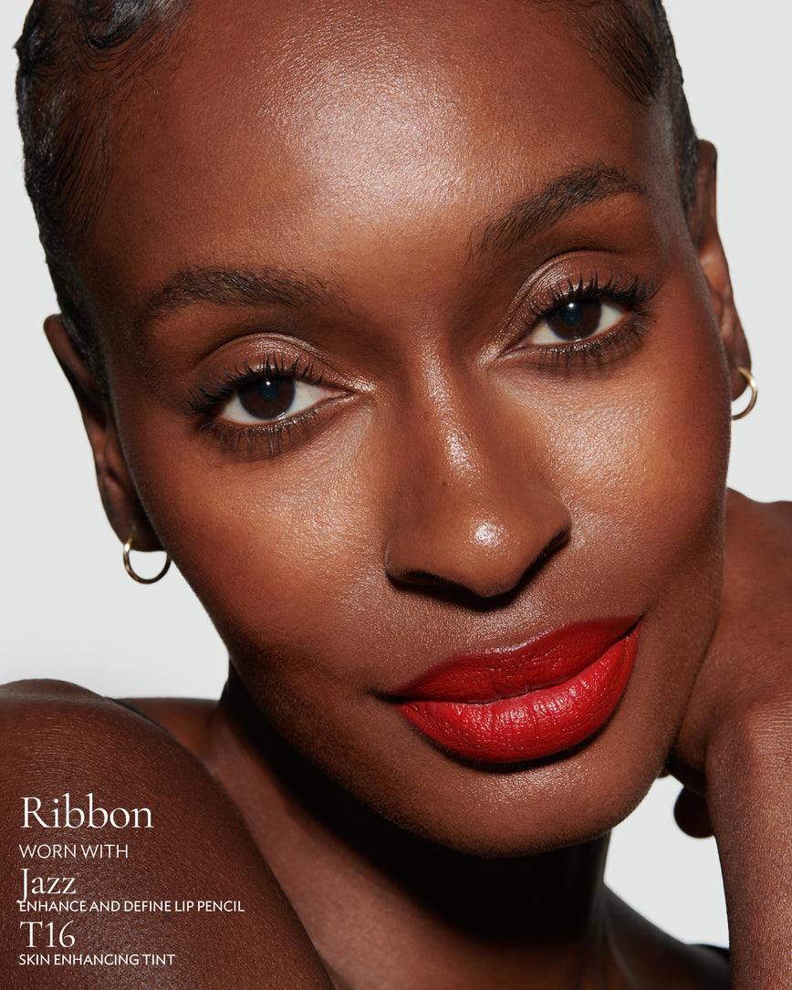 Close-up of a woman with a grey background, focusing on the lip colour - Pillar-box red Ribbon, with notes on the Jazz Enhance and Define Pencil and T16 Skin Enhancing Tint
