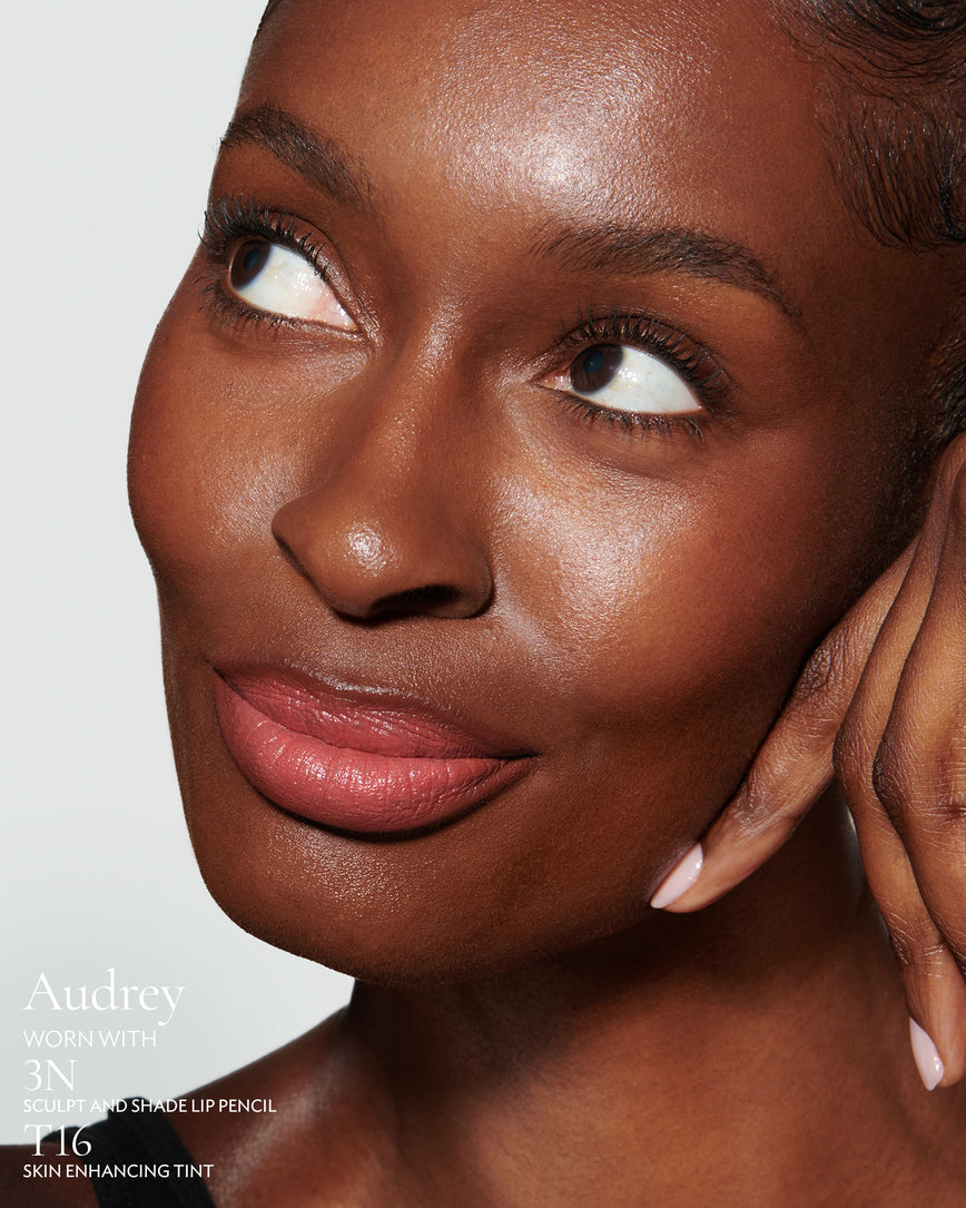 Close-up of a woman with a grey background, focusing on the lip colour - salmon pink Audrey, with notes on the 3N Sculpt and Shade Pencil and T16 Skin Enhancing Tint