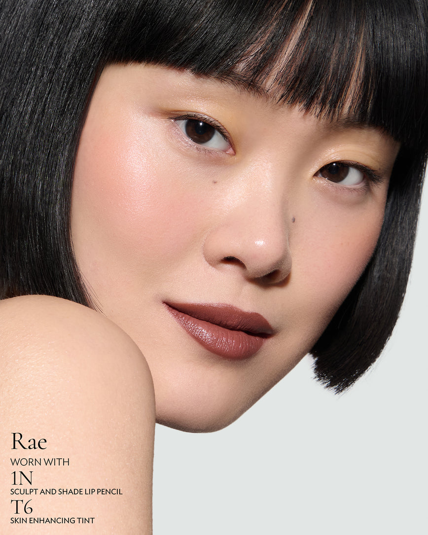 Close-up of a woman with a grey background, focusing on the lip colour - red-chocolate Rae, with notes on the 1N Sculpt and Shade Pencil and T6 Skin Enhancing Tint