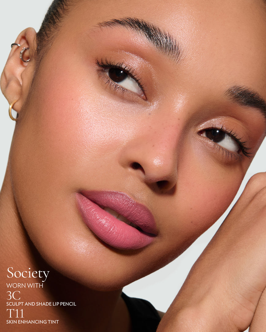 Close-up of a woman with a grey background, focusing on the lip colour - warm pink Society, with notes on the 3C Sculpt and Shade Pencil and T11 Skin Enhancing Tint