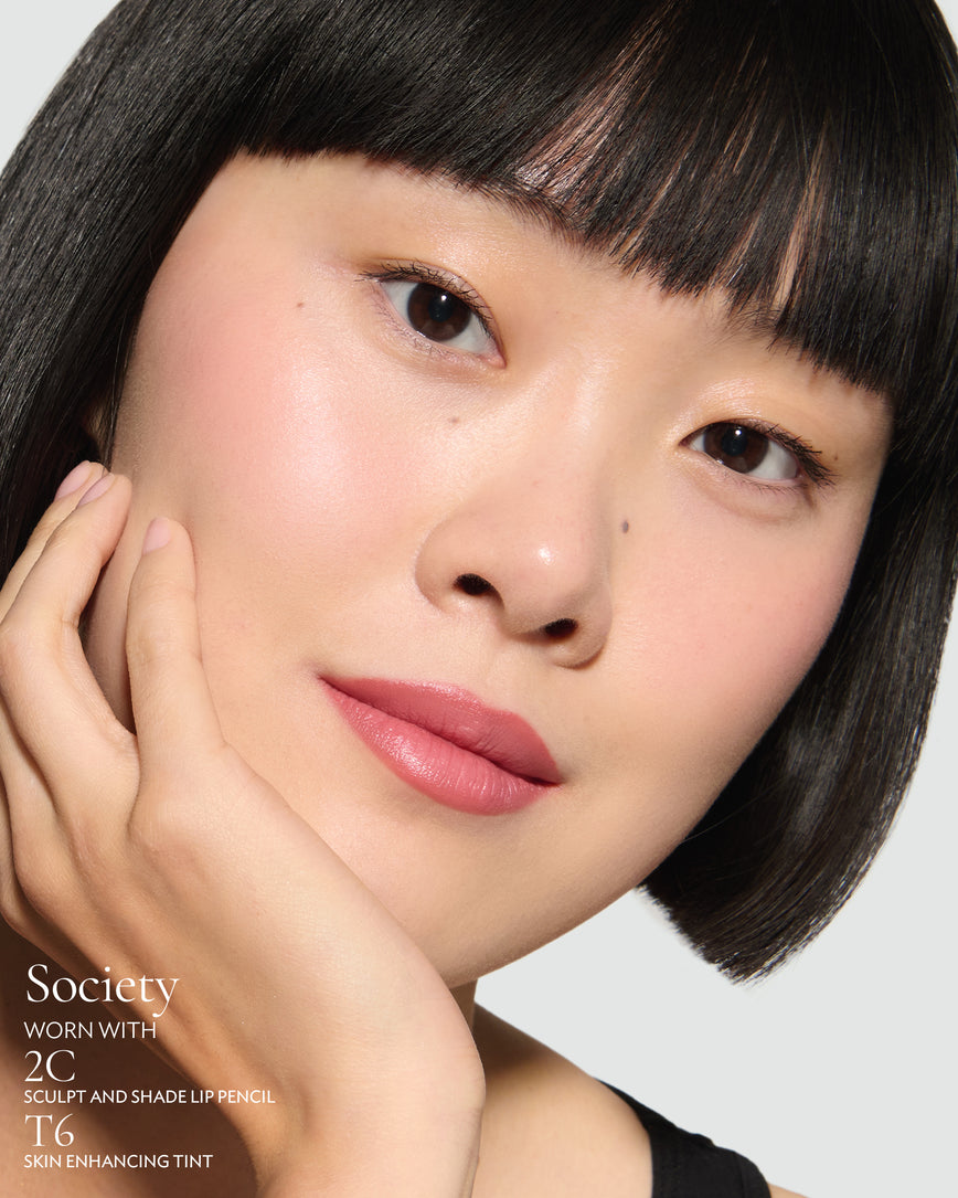 Close-up of a woman with a grey background, focusing on the lip colour - warm pink Society, with notes on the 2C Sculpt and Shade Pencil and T6 Skin Enhancing Tint