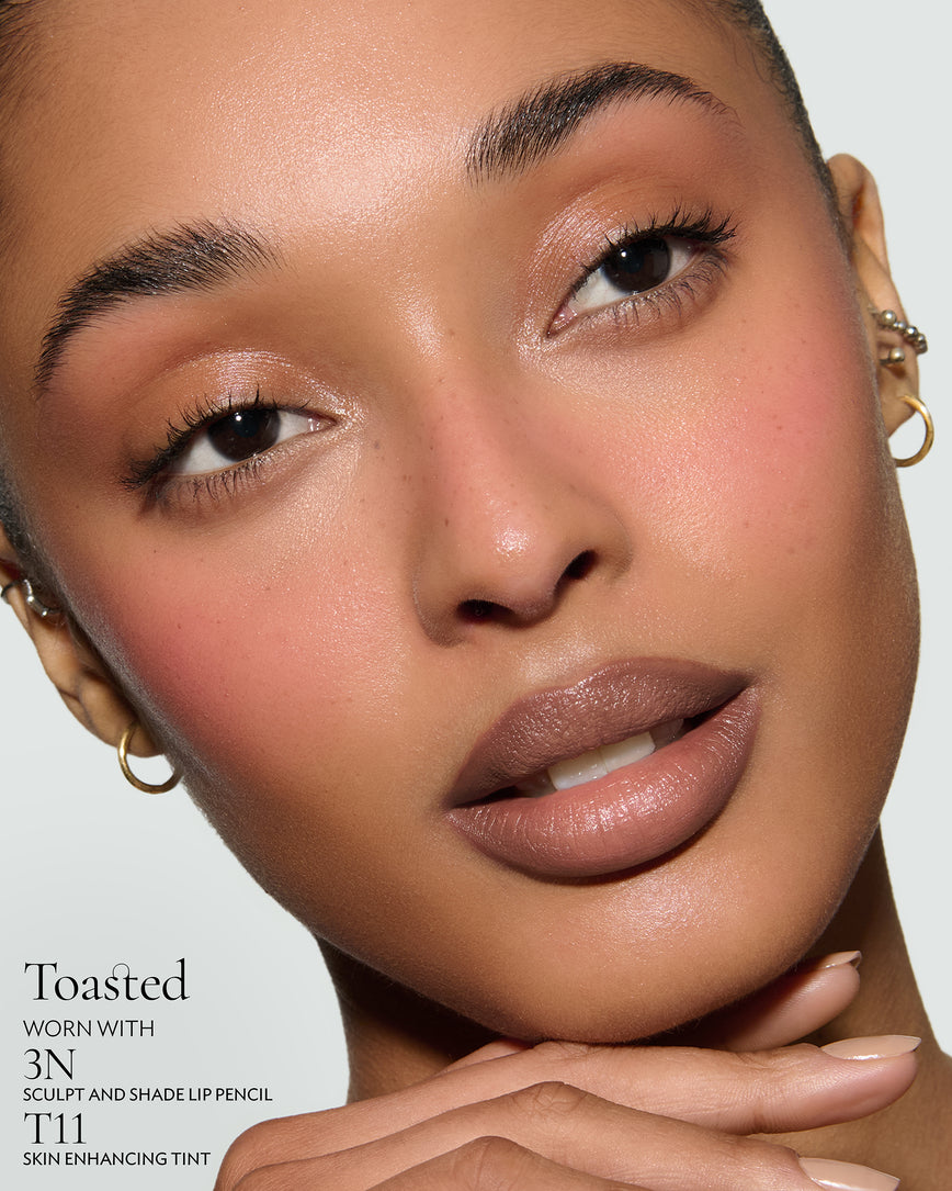 Close-up of a woman with a grey background, focusing on the lip colour - neutral-beige Toasted, with notes on the 3N Sculpt and Shade Pencil and T11 Skin Enhancing Tint