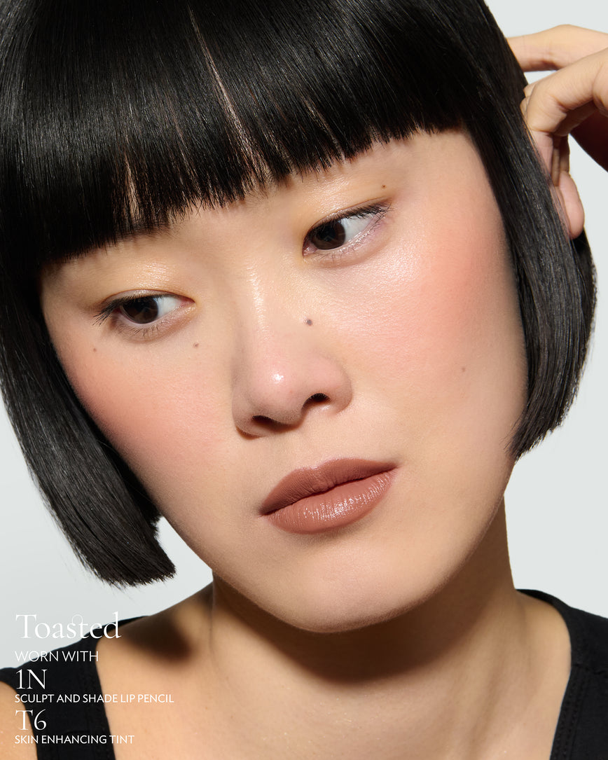 Close-up of a woman with a grey background, focusing on the lip colour - neutral-beige Toasted, with notes on the 1N Sculpt and Shade Pencil and T6 Skin Enhancing Tint 