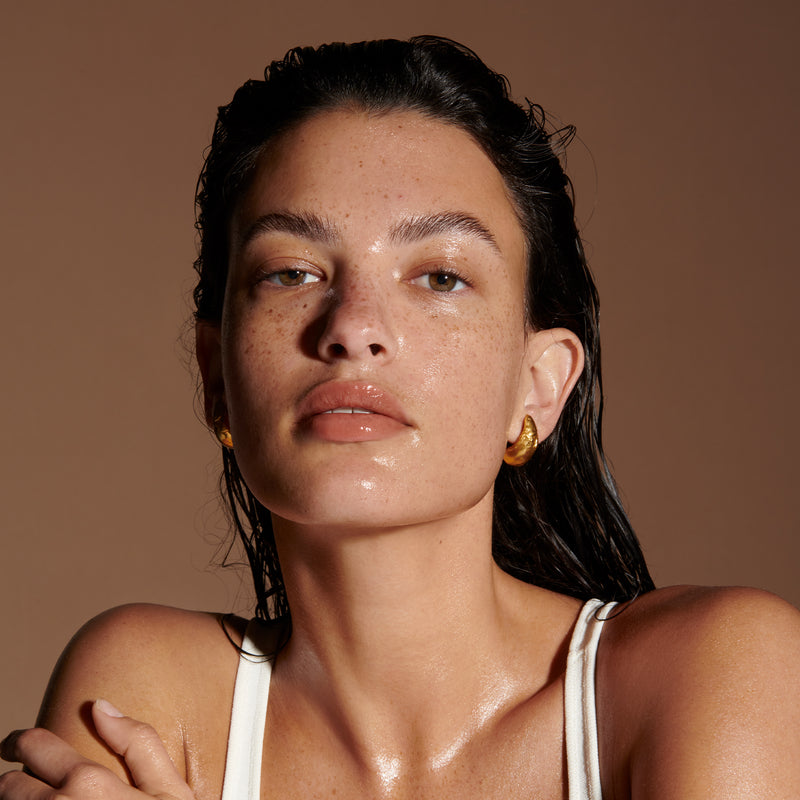The head and shoulders of a model with glistening, tan skin. Their dark hair is wet and pushed back from their face and goes down their back. They have a natural makeup look on with a shine on their eyelids, nose, lips and chin.