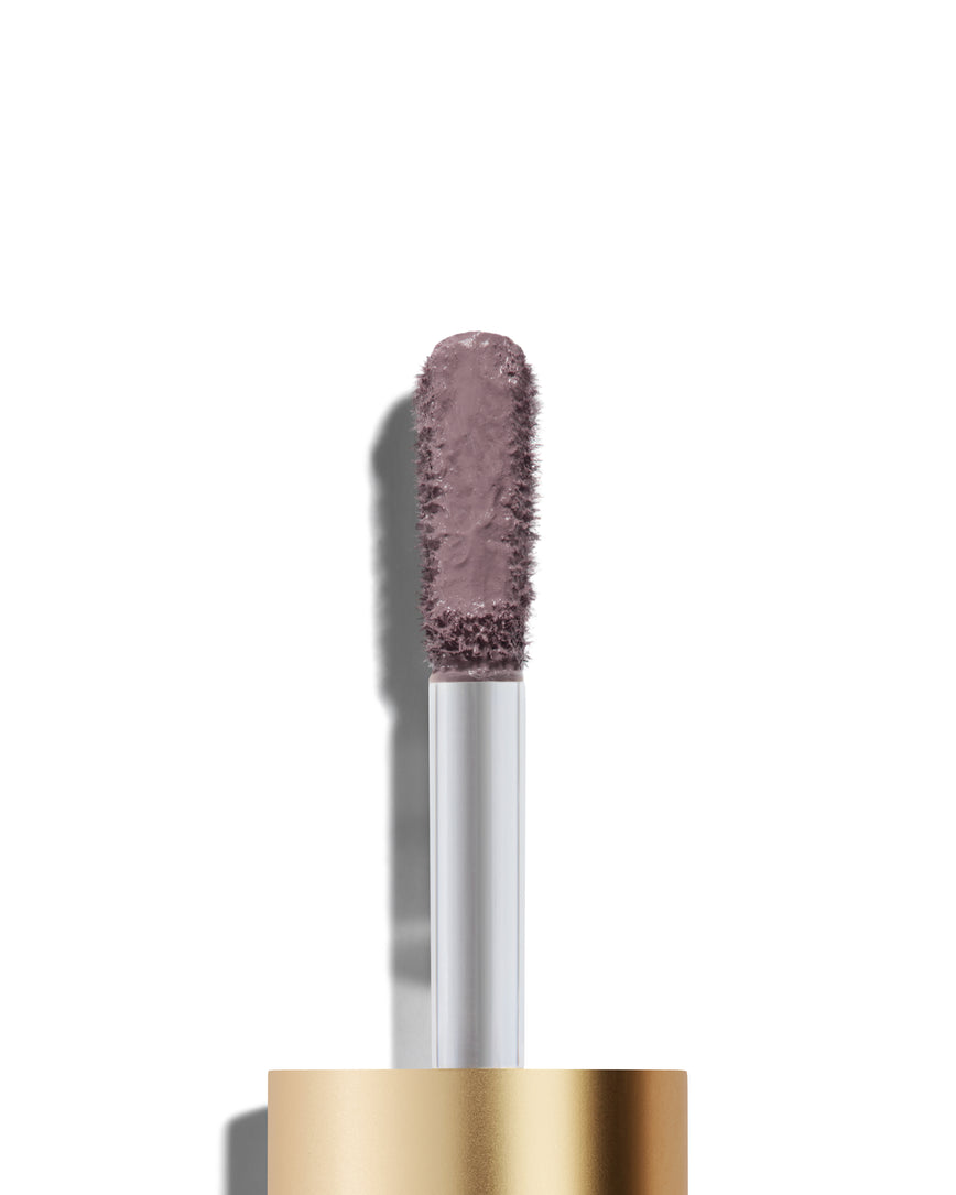 The end of a gold cap containing an applicator wand with a Maude Liquid Silk eyeshadow on it, against a white background.