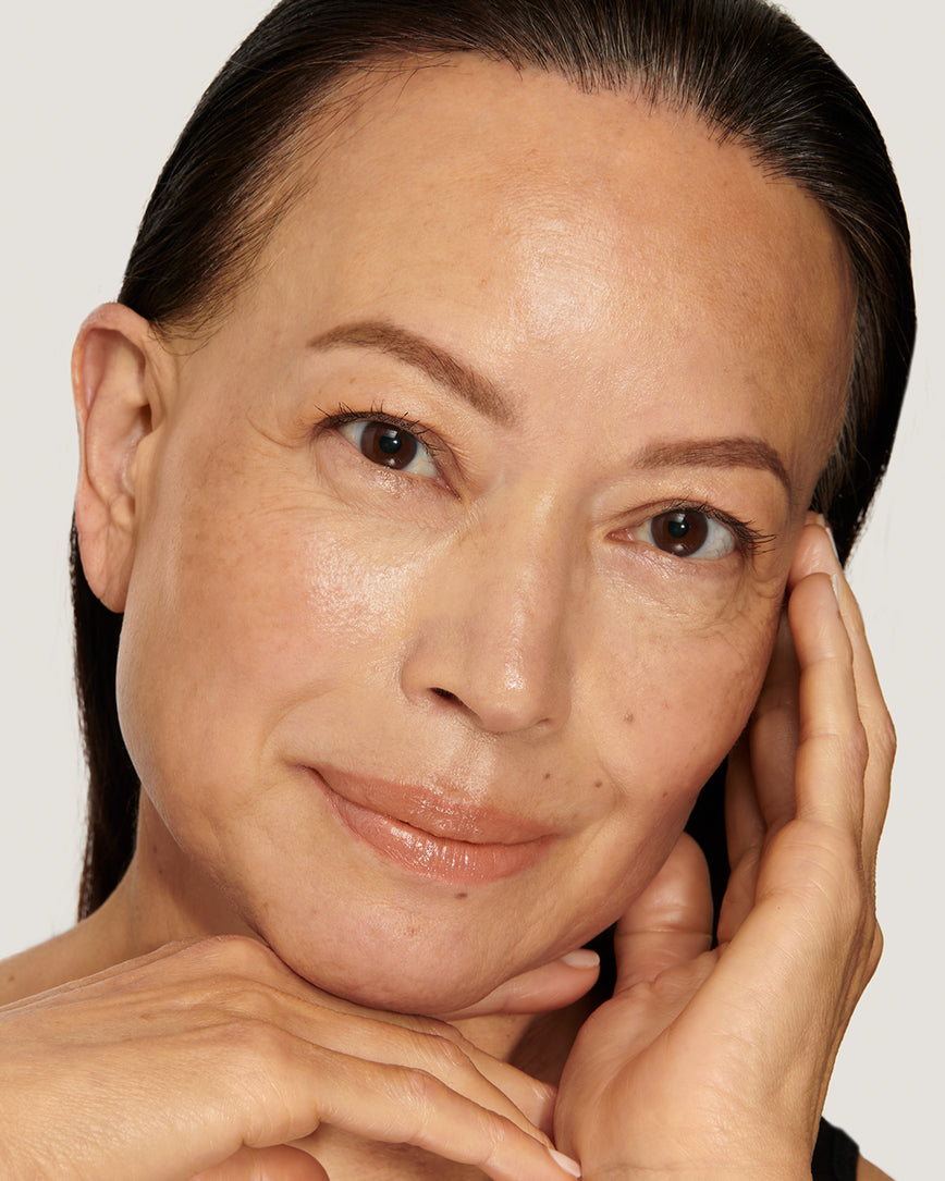 Model with a warm smile, touching her face against a neutral background, wearing Meet Cute Baume Embrace