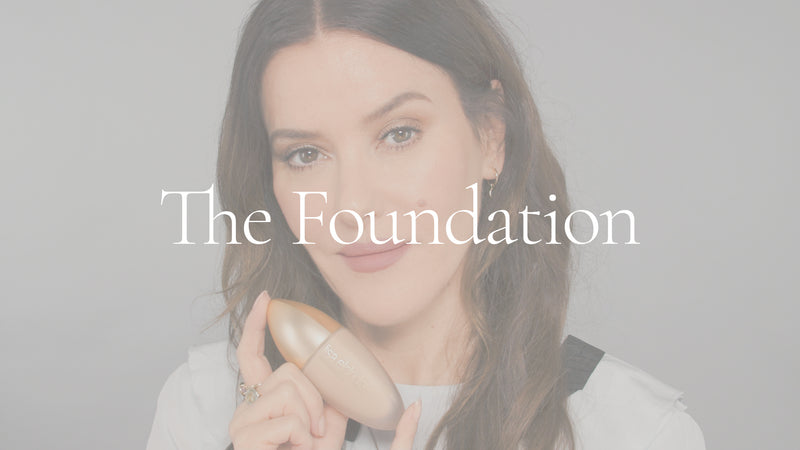 Poster image with white overlay for a video. Lisa Eldridge is in front of a grey background. She holds an elongated matte glass bottle with a gold lid. On top of the image is the text, “The Foundation”.