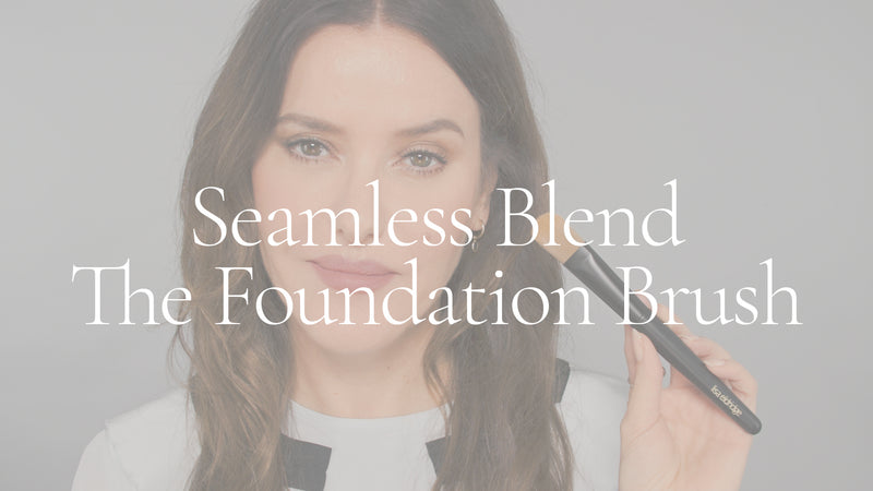 Poster image with white overlay for a video. Lisa Eldridge is in front of a grey background. She holds a foundation brush with a flat, rounded brush and black handle. On top of the image is the text, “Seamless Blend The Foundation Brush”.