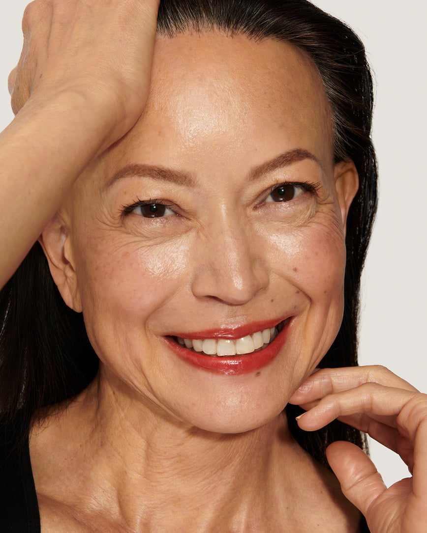Model with a warm smile, touching her hair and face against a neutral background, wearing Red Curve Baume Embrace