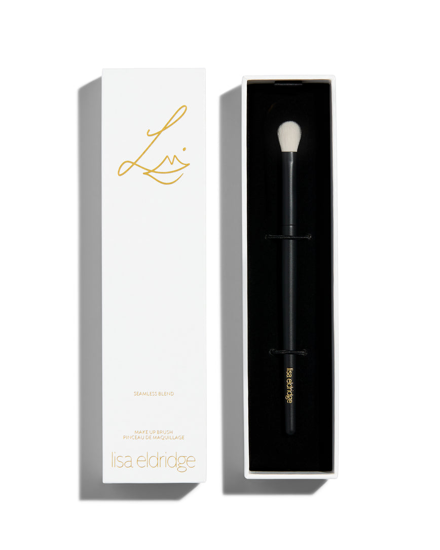 An open rectangular white box with black interior. Inside is a foundation brush with a long black handle and a short rounded tip with white bristles. The lid of the box sits next to the bottom and has the Lisa Eldridge logo in gold as well as the text, ‘SEAMLESS BLEND MAKE UP BRUSH PINCEAU DE MAQUILLAGE lisa eldridge’ 