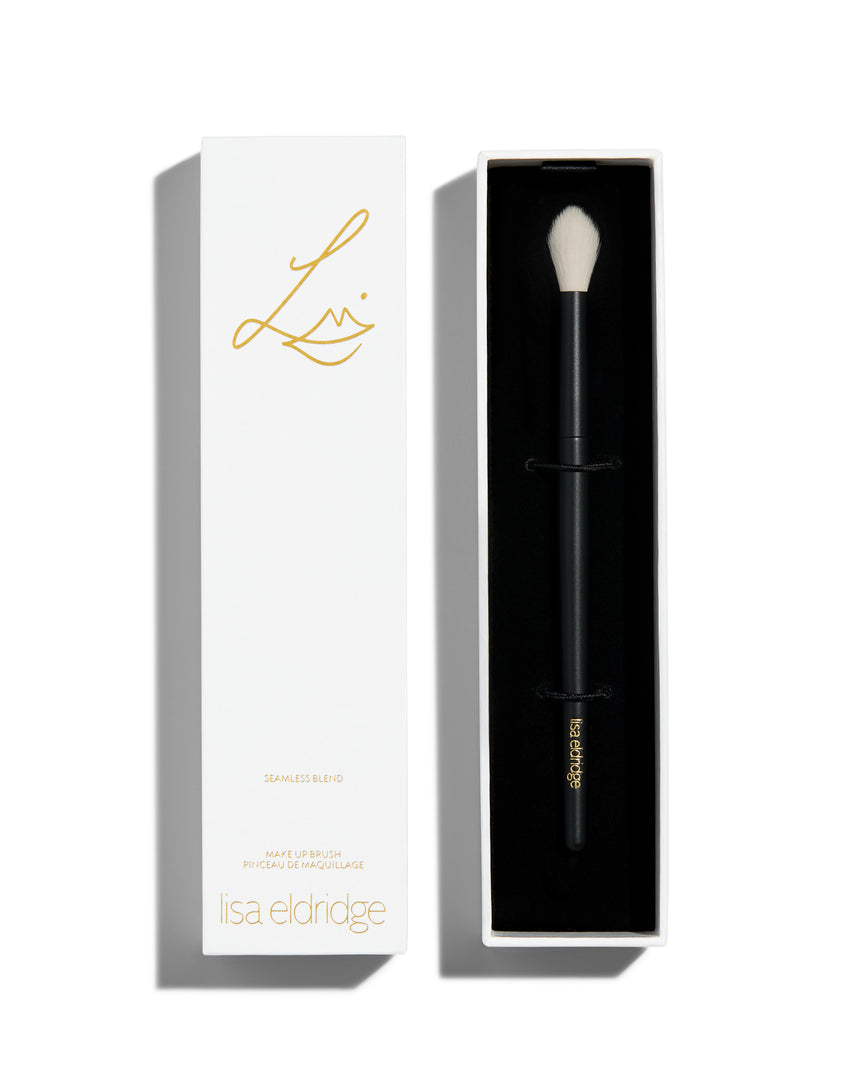 An open rectangular white box with black interior. Inside is a foundation brush with a long black handle and a rounded tip with long, tapered white bristles. The lid of the box sits next to the bottom and has the Lisa Eldridge logo in gold as well as the text, ‘SEAMLESS BLEND MAKE UP BRUSH PINCEAU DE MAQUILLAGE lisa eldridge’ 
