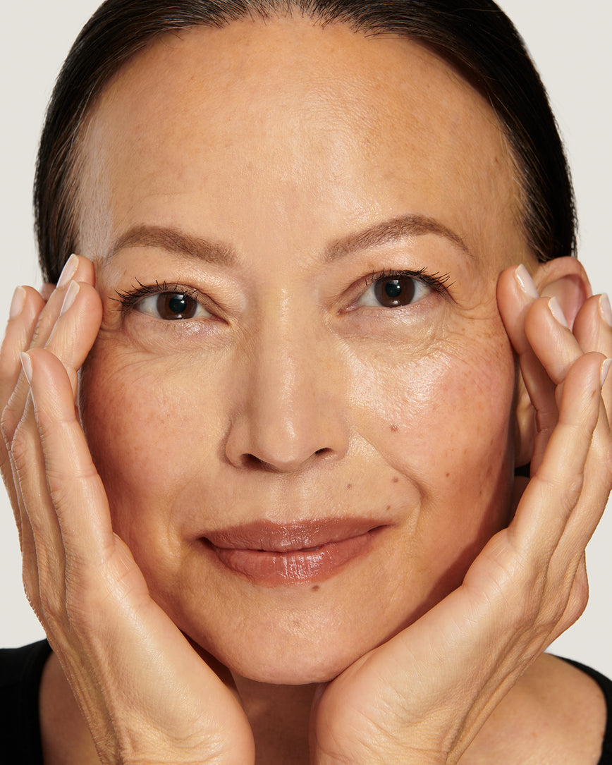 Model with a warm smile, touching her face against a neutral background, wearing Sweet Fig Baume Embrace