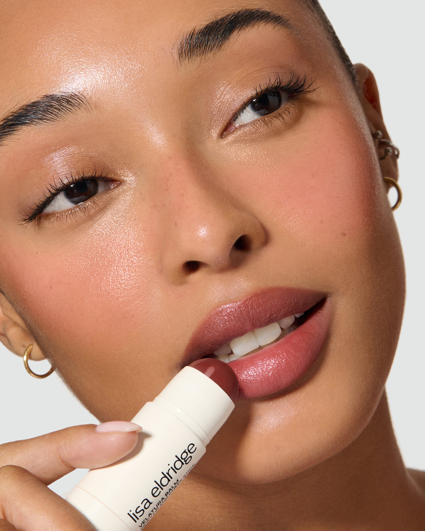 Extreme close-up of a model's face with a neutral background, wearing Cinnamon Bun Velatura Balm on lip and cheeks and applying the product to her lips