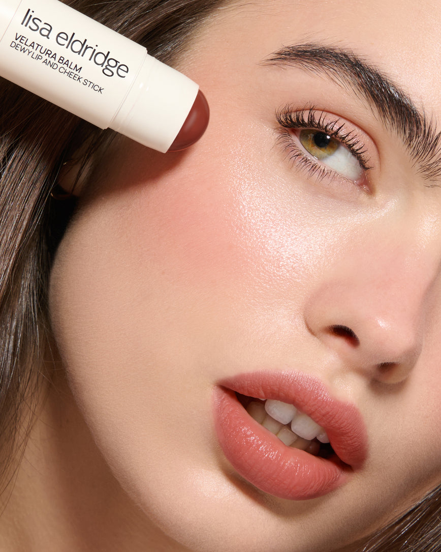 Extreme close-up of a model's face with a neutral background, wearing Cinnamon Bun Velatura Balm on lip and cheeks and applying the product to her cheek