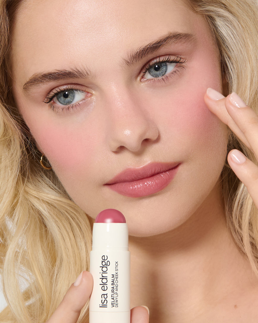 Close-up of a model's face with a neutral background, wearing Rose Madder Velatura Balm on lip and cheeks and applying the product to her lips