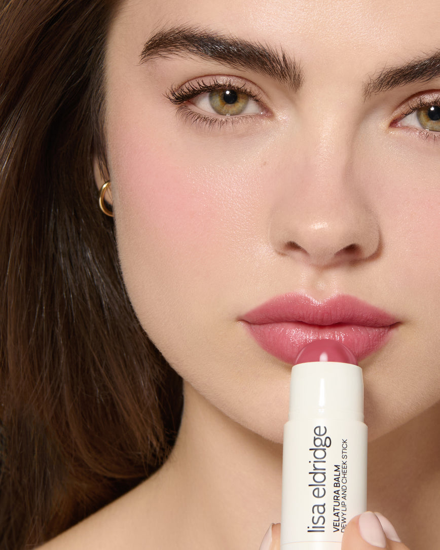Close-up of a model's face with a neutral background, wearing Rose Madder Velatura Balm on lip and cheeks and applying the product to her lips
