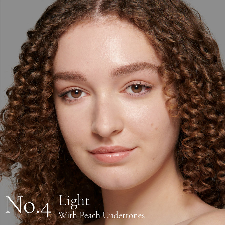 Lisa Eldridge Seamless Skin Foundation Pictured on Model. A young model with light skin and peach undertones, brown eyes and brown hair and eyebrows. The text says ' No.4 Light With Peach Undertones'