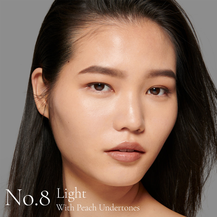 Lisa Eldridge Seamless Skin Foundation Pictured on Model. A young model with light skin and peach undertones, brown eyes and straight black hair. The text says ' No.8 Light With Peach Undertones’