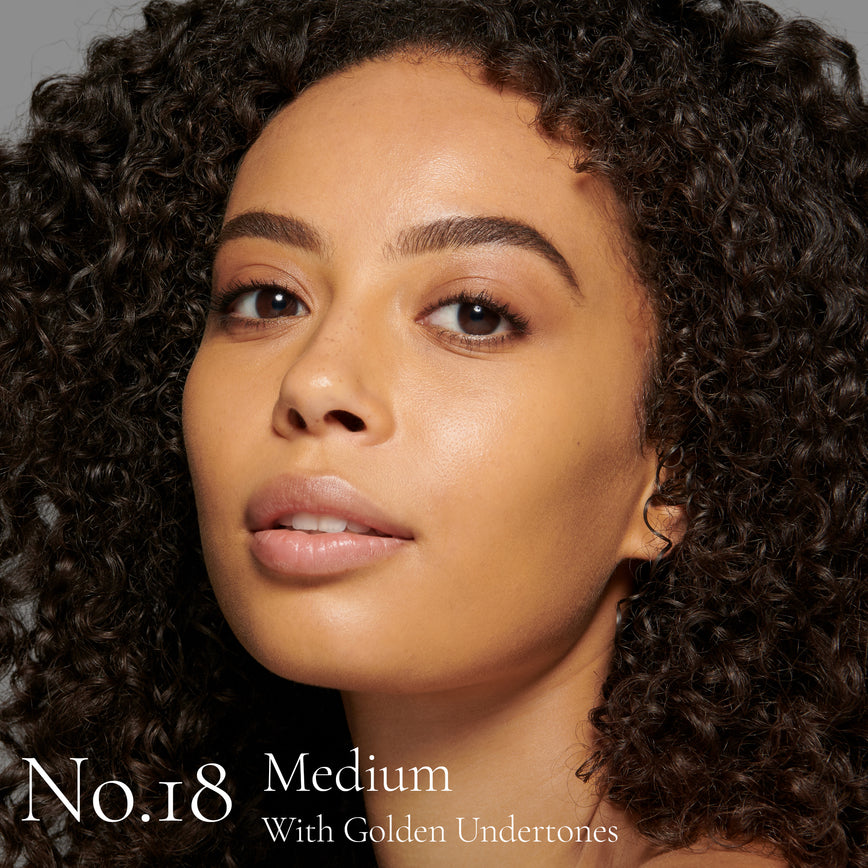 Lisa Eldridge Seamless Skin Foundation Pictured on Model. The model has medium skin and golden undertones, brown eyes and curly black hair. The text says ‘No.18 Medium With Golden Undertones’