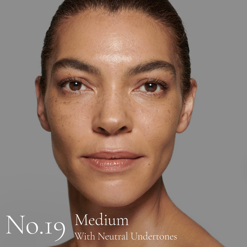 Lisa Eldridge Seamless Skin Foundation Pictured on Model. The model has medium skin and neutral undertones, brown eyes and dark brown hair that is tied back. The text says ‘No.19 Medium With Neutral Undertones’