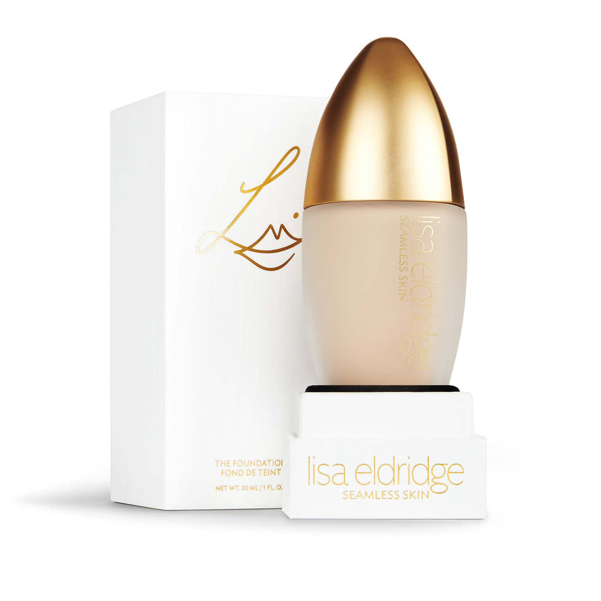 Lisa Eldridge Seamless Skin Foundation in a matte glass bottle with a gold lid. Behind the product is the box it comes packaged in. It is a white rectangular box with the Lisa Eldridge logo in gold.