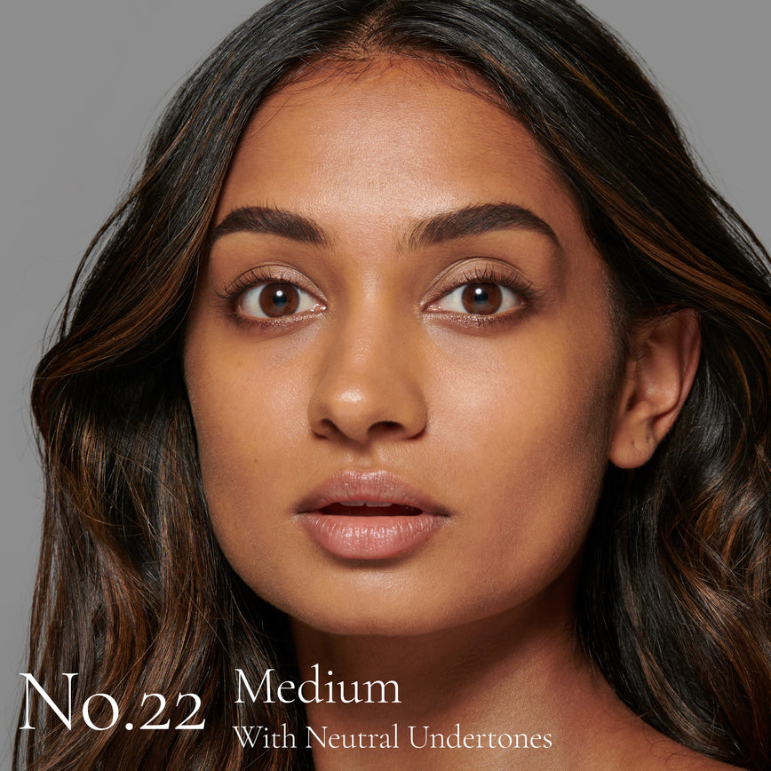 Lisa Eldridge Seamless Skin Foundation Pictured on Model. The model has medium skin and neutral undertones, brown eyes and slightly wavy black hair with some reddish highlights. The text says ‘No.22 Medium With Neutral Undertones’