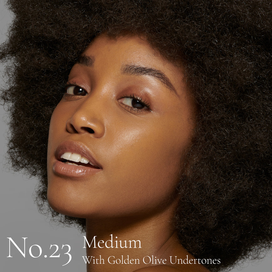 Lisa Eldridge Seamless Skin Foundation Pictured on Model. The model has medium skin and golden olive undertones, brown eyes and curly black hair. The text says ‘No.23 Medium With Golden Olive Undertones’