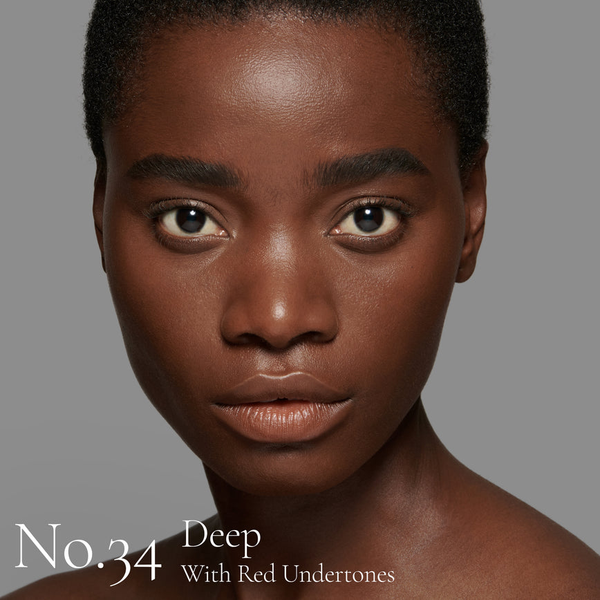 Lisa Eldridge Seamless Skin Foundation pictured on model. The model has deep skin and red undertones, brown eyes and black hair. The text says ‘No.34 Deep With Red Undertones’