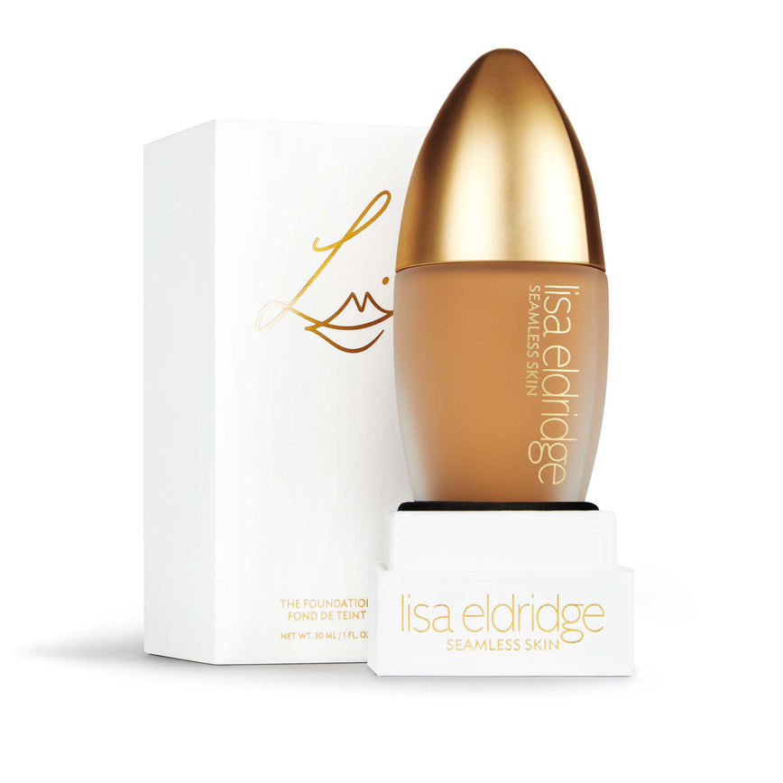Lisa Eldridge Seamless Skin Foundation in a matte glass bottle with a gold lid. Behind the product is the box it comes packaged in. It is a white rectangular box with the Lisa eldridge logo in gold.