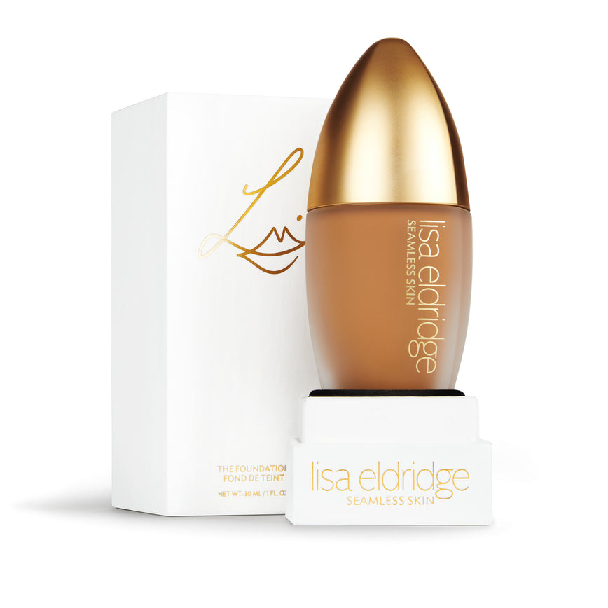 Lisa Eldridge Seamless Skin Foundation in a matte glass bottle with a gold lid. Behind the product is the box it comes packaged in. It is a white rectangular box with the Lisa eldridge logo in gold.