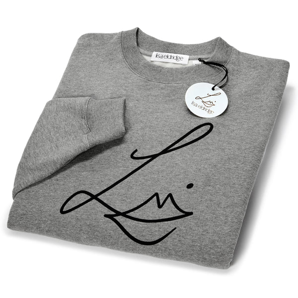 The Lisa Eldridge Studio Sweatshirt in grey folded up. The crewneck and the cuffs have a fine rib. The Lisa Eldridge logo is large on the front. There is a label that says “lisa eldridge’ sewn on the inside. Attached to the size label is a circular card tag on a black ribbon with the Lisa Eldridge logo in gold.