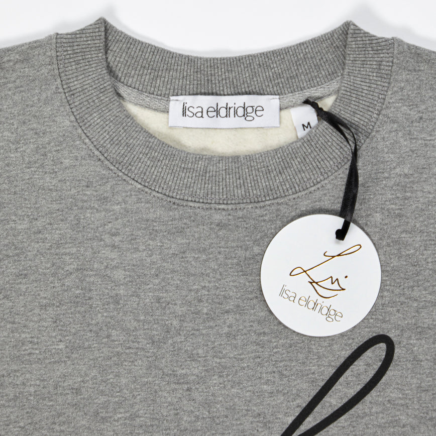 Close-up of the the Lisa Eldridge Studio Sweatshirt in grey. The crewneck has a fine rib. There is a label that says “lisa eldridge’ sewn on the inside. Attached to the size label is a circular card tag on a black ribbon with the Lisa Eldridge logo in gold.